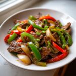 A close-up of vibrant pepper steak featuring strips of beef, green bell peppers, red peppers, and onions coated in a savory sauce.
