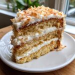 A tall slice of three-layer coconut cake with white frosting and toasted coconut flakes on top.