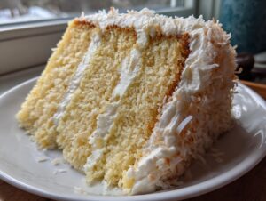 Close-up of a moist slice of 3-layer coconut cake with white frosting and toasted coconut flakes.