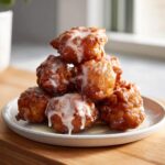 A stack of golden brown, freshly fried apple fritter pieces drizzled generously with white glaze.