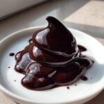 A close-up of thick, glossy, dark chocolate ganache piped into a swirl shape on a white plate.