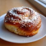 A close-up of a freshly baked, golden brown pan de Mallorca Puerto Rican sweet roll dusted heavily with powdered sugar.
