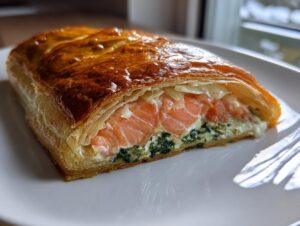 Close-up of a slice of salmon wellington showing pink salmon, creamy spinach filling, and golden puff pastry.