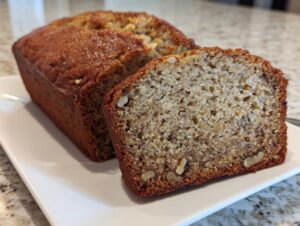 A freshly baked loaf of greek yogurt banana bread, with one thick slice cut and resting beside it on a white plate.