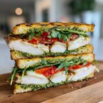 A tall, stacked cross-section of a grilled chicken pesto sandwich showing layers of chicken, melted cheese, tomato, and arugula on toasted bread.