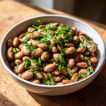 Close-up of a white bowl filled with moist, cooked healthy pinto beans garnished generously with chopped green parsley.