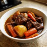Close-up of a white bowl filled with rich, dark beef stew, tender chunks of beef, carrots, and potatoes.