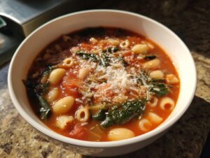 A close-up view of a steaming bowl of minestrone soup, featuring pasta, white beans, tomatoes, greens, and topped with grated Parmesan cheese.