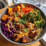 A vibrant, colorful buddha bowl filled with quinoa, crispy tofu, roasted sweet potatoes, broccoli, red cabbage, and drizzled with peanut sauce.