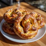 A stack of golden-brown, freshly baked high-protein high-fiber pretzels topped with coarse salt on a white plate.