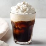A glass of iced coffee topped with a layer of light brown cold foam and whipped cream.
