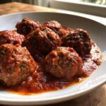 A close-up view of several juicy Italian meatballs simmering in a rich, thick red tomato sauce in a white bowl.