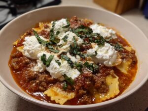 A close-up of a bowl of rich lasagna soup featuring pasta, meat sauce, dollops of ricotta, and fresh basil.