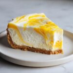 A close-up of a single slice of lemon curd cheesecake showing a graham cracker crust and a white filling swirled with bright yellow lemon curd.