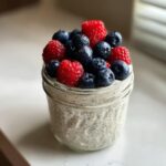 Close-up of a low calorie chia pudding snack in a glass jar topped generously with fresh blueberries and raspberries.