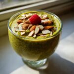 A glass of bright green matcha chia pudding topped with sliced almonds and a single fresh raspberry.
