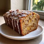A moist apple dapple cake loaf drizzled with white glaze and topped with pecans, sitting on a white plate.
