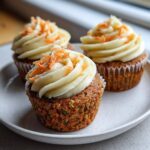 Close-up of three moist carrot cake cupcakes topped with thick cream cheese frosting and shredded carrots.