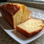 A golden-brown loaf of cream cheese pound cake with one thick slice cut and resting beside it on a white platter.