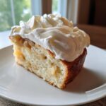 A slice of moist German apple cake studded with apple pieces and topped generously with swirls of white cream.