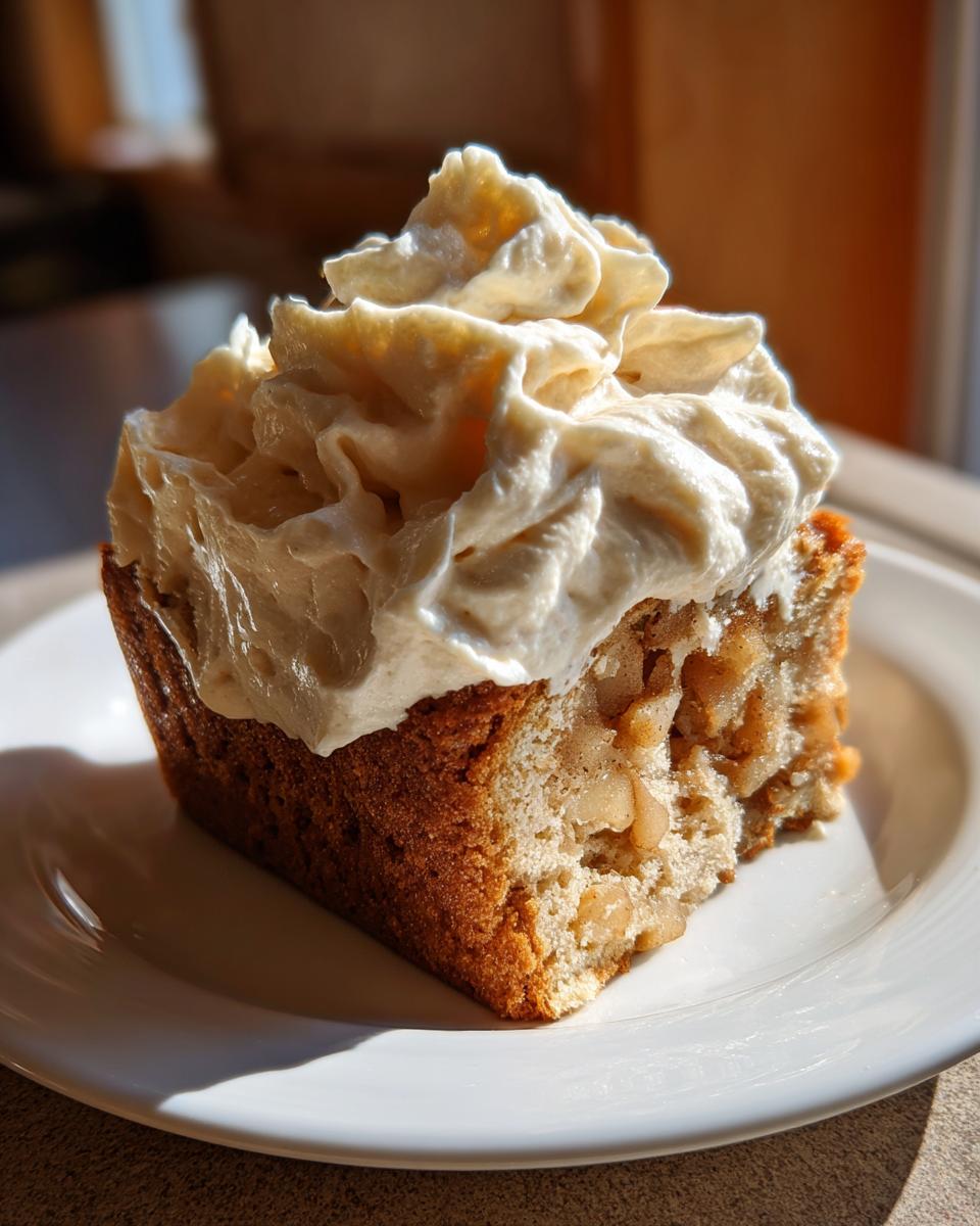 A close-up of a slice of moist German apple cake topped generously with fluffy amaretto cream.