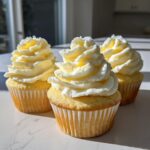 Three moist lemon cupcakes topped with swirls of pale yellow and white frosting, sitting on a light surface.