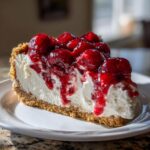 A close-up of a perfect slice of no-bake cherry cheesecake featuring a graham cracker crust and thick cherry topping.