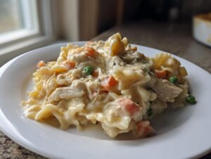 A close-up of a serving of creamy chicken pot pie pasta featuring bowtie noodles, shredded chicken, peas, and carrots on a white plate.