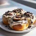 Four freshly baked Oreo cinnamon rolls covered in thick white frosting and crushed Oreo pieces, sitting on a white plate.
