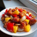 A close-up of glossy, saucy pineapple chicken stir-fry pieces mixed with chunks of pineapple, red bell pepper, and onion.
