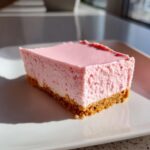 A close-up of a thick, square pink birthday cheesecake slice with a visible graham cracker crust.