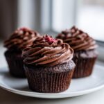 Three rich, dark chocolate cupcakes topped with swirled chocolate frosting and a sprinkle of raspberry on top, ready to eat.