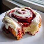 A close-up photo of a single, decadent red velvet cinnamon rolls topped with thick cream cheese frosting, with a bite taken out.