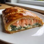 Close-up of a slice of salmon wellington showing flaky golden pastry wrapped around pink salmon and creamy spinach filling.