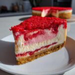 A close-up slice of strawberry crunch cheesecake featuring layers of creamy filling, strawberry swirl, and a bright red crumb topping.