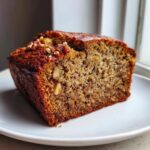 A thick slice of moist sugar free banana bread topped with walnuts, resting on a white plate.