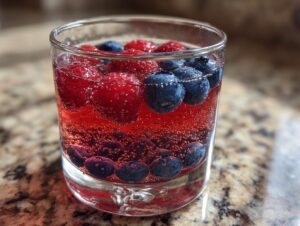 Close-up of a glass containing red, bubbly patriotic punch garnished with fresh raspberries and blueberries.