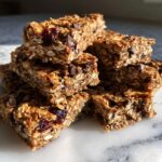 A close-up stack of chewy no bake granola bars featuring oats, dried cranberries, and chocolate chips on a white marble surface.