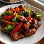 Close-up of glossy teriyaki chicken stir fry featuring chicken pieces, bright green broccoli, red peppers, and carrots.