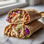 Two halves of a thai peanut chicken wrap stacked, showing shredded chicken, colorful slaw, and peanut sauce drizzling down.