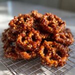 A close-up of a pile of crunchy, glazed toffee pretzels coated in a sticky, nutty toffee mixture, cooling on a wire rack.