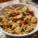 A close-up serving of creamy Tuscan chicken orzo pasta mixed with sun-dried tomatoes and spinach in a white bowl.