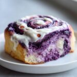 A close-up of a single, fluffy ube cinnamon roll topped with white cream cheese icing, showing the vibrant purple ube swirl.