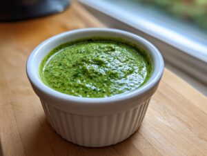 A close-up of vibrant, bright green cilantro lime sauce resting in a small white fluted ramekin on a wooden surface.