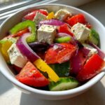 Close-up of a fresh greek salad in a white bowl featuring large chunks of tomato, cucumber, red onion, bell peppers, Kalamata olives, and feta cheese.