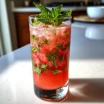 A tall glass filled with a vibrant watermelon mint mojito, garnished with fresh mint sprigs and ice.