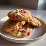 A stack of four freshly baked white chocolate raspberry cookies featuring visible white chocolate chips and bright red raspberry pieces.