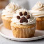 Close-up of a vanilla Dog Mom Day cupcake topped with white frosting and a chocolate sprinkle paw print.