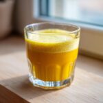 A vibrant, foamy Ginger lemon turmeric shot served in a small, faceted glass on a wooden surface.