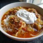 Close-up of a white bowl filled with Golumpki soup, featuring cabbage, meatball, rice, and a large dollop of sour cream.